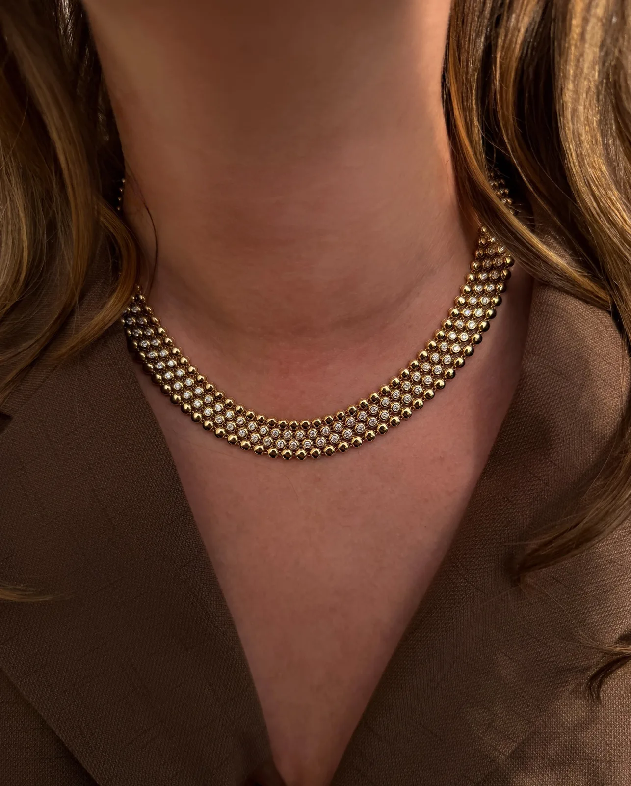 A wide yellow gold collar necklace with rows of bezel-set diamonds, worn against a cocoa blazer